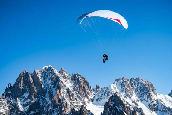 Un parapentiste survole des montagnes enneigées sous un ciel bleu.