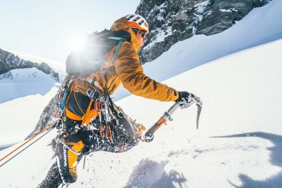 Alpiniste en orange grimpant une pente enneigée avec un piolet.