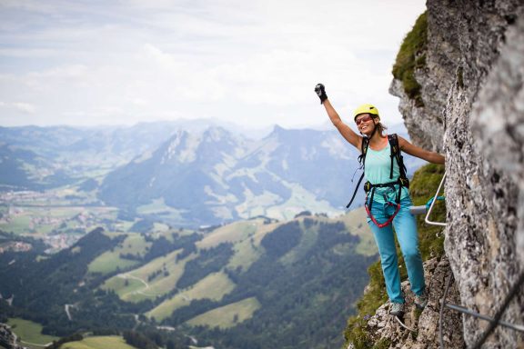 Jeune femme escaladant une paroi rocheuse, main levée en signe de victoire, paysage alpin en arrière-plan.
