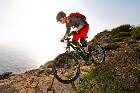 Cycliste en action sur un sentier escarpé, bord de mer, sous un soleil éclatant.