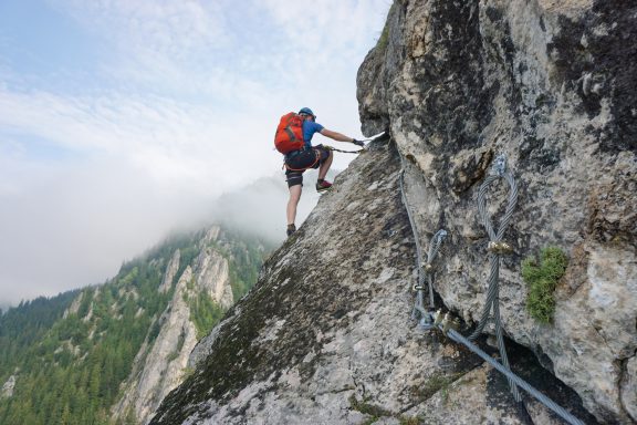 Un homme grimpe une paroi rocheuse avec un harnais et un sac à dos dans un paysage montagneux.